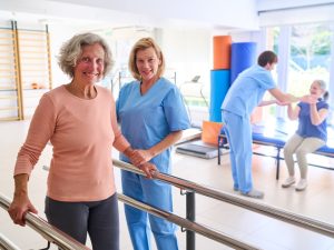A,Senior,Woman,Walks,With,Parallel,Bars,Assisted,By,A Senior woman walking with assistance in a rehabilitation facility